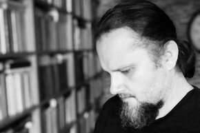 Przemysław Karbowski (mężczyzna z brodą oraz długimi włosami spiętymi w kucyk) czyta książkę. W tle półka z książkami. 