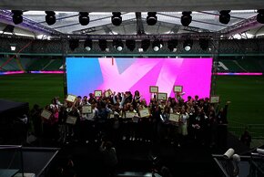 Grupa kilkudziesięciu młodych osób stoi na scenie stadionu piłkarskiego, trzymając dyplomy i radośnie pozując do zdjęcia. Za nimi znajduje się duży, kolorowy ekran LED w odcieniach różu i błękitu. Uczestnicy mają przypięte identyfikatory i uśmiechnięte twarze, a z przodu kilku fotografów uwiecznia chwilę. W tle widoczne są trybuny stadionowe z zielonymi krzesełkami.