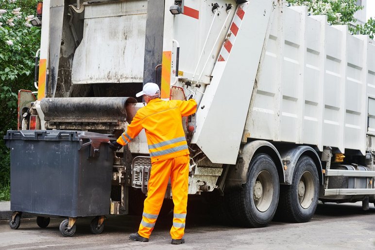 Śmieciarka odbiera odpady z pojemnika, przed nią pracownik ubrany w pomarańczowy strój roboczy. Stock images by Depositphotos. 