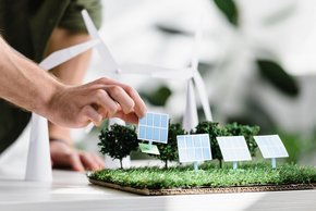 Zdjęcie ilustracyjne. Ręka wkłada panel fotowoltaiczny w model, gdzie są już drzewa i wiatraki. Stock images by Depositphotos. 