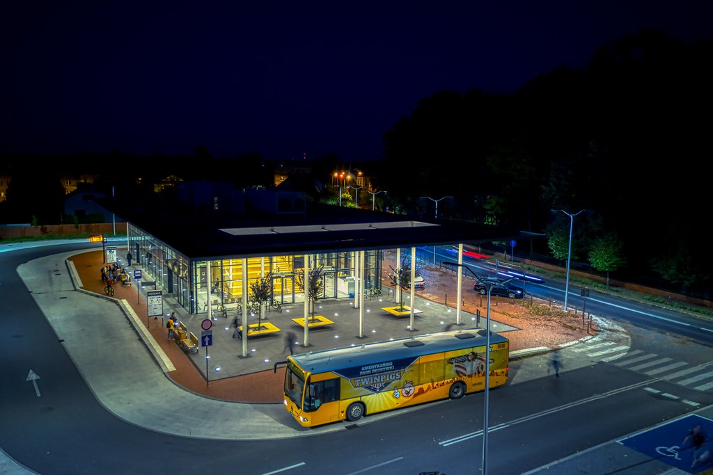 Nocne zdjęcie rozświetlonego centrum przesiadkowego, na pierwszym planie autobus BKM oklejony w barwach parku rozrywki Twinpigs. 