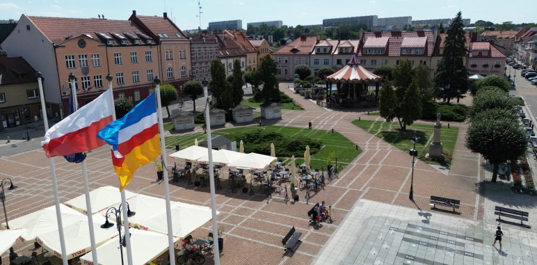 Widok z lotu ptaka na rynek w Żorach w słoneczny dzień. W centralnej części widoczne są stoliki z parasolami restauracyjnymi oraz karuzela. Na pierwszym planie powiewają flagi: Polski i Żor. W tle zabytkowe kamienice, zieleń i alejki spacerowe.