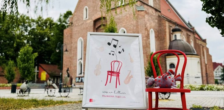 Czerwone krzesło z maskotką „Kulturkiem” stoi obok plakatu promującego wydarzenie „Czas na kulturę. Posłuchaj nas. Znajdź czas.” W tle widoczny zabytkowy kościół pw. św. św. Apostołów Filipa i Jakuba w Żorach.